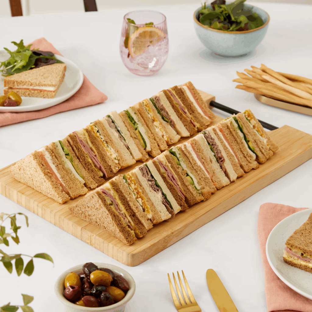 Gourmet Grocer Sandwich platter on wooden board with pot of olives, plated sandwiches, side salad and bread sticks